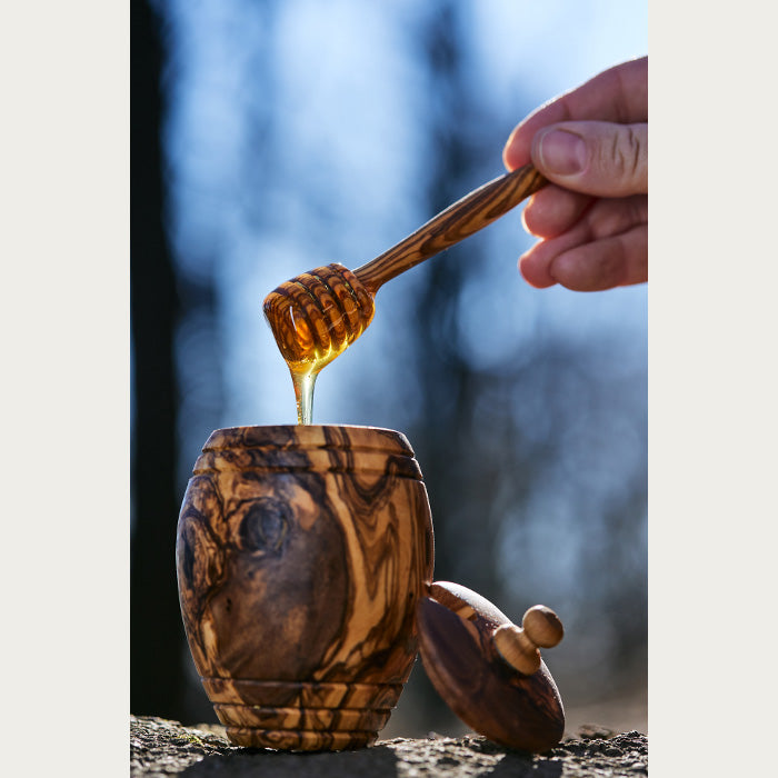 Ruka drži kašiku za med iznad drvene posude.