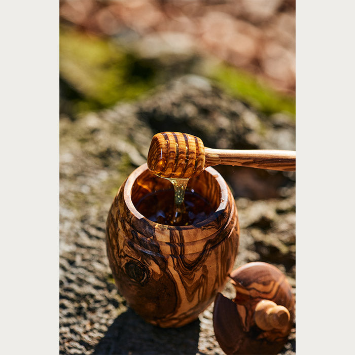 Drvena posuda puna meda i kašika sa koje se preliva med, na kamenu.