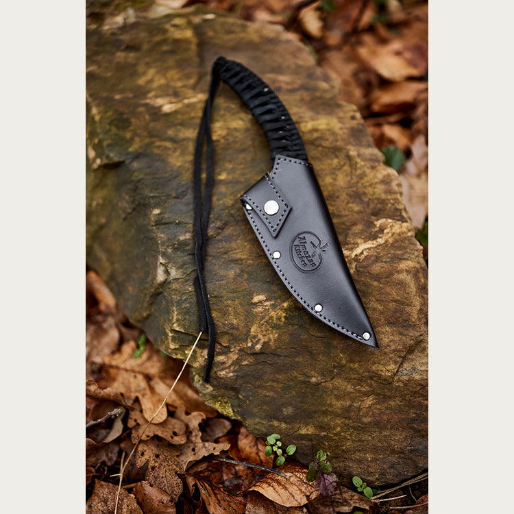 Almazan Kitchen Predator Nož u crnoj kožnoj futroli, položen na kamen u šumi. 