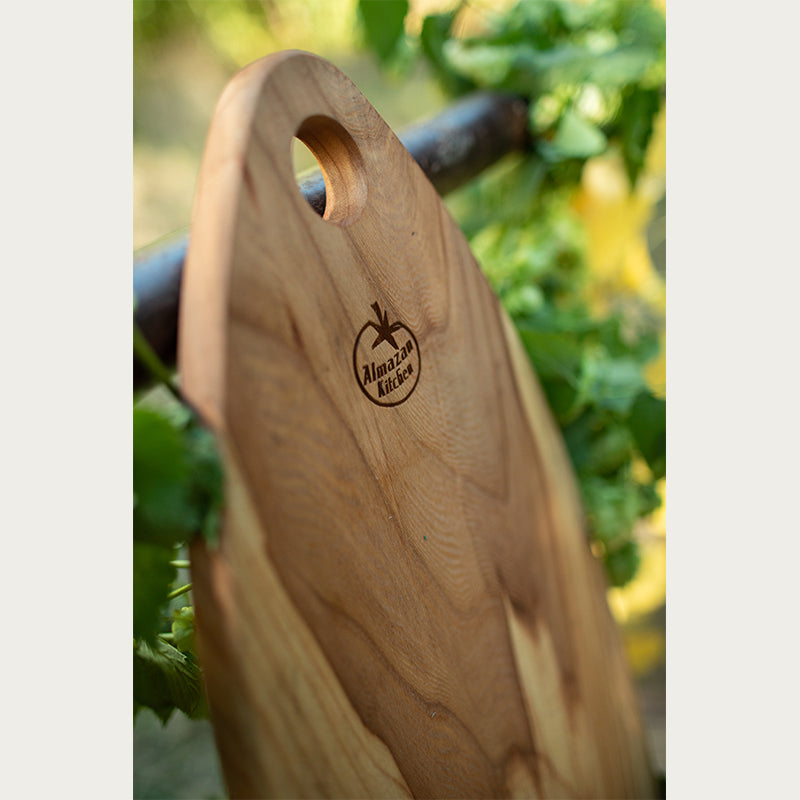 Almazan Kitchen daska od orahovog drveta naslonjena na ogradu obraslu lišćem sa logom u fokusu