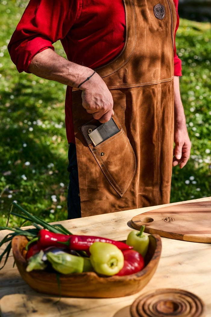 Čovek izvlači nož iz džepa na kožnoj kuvarskoj kecelji i sprema se da seče povrće koje je ispred njega. 