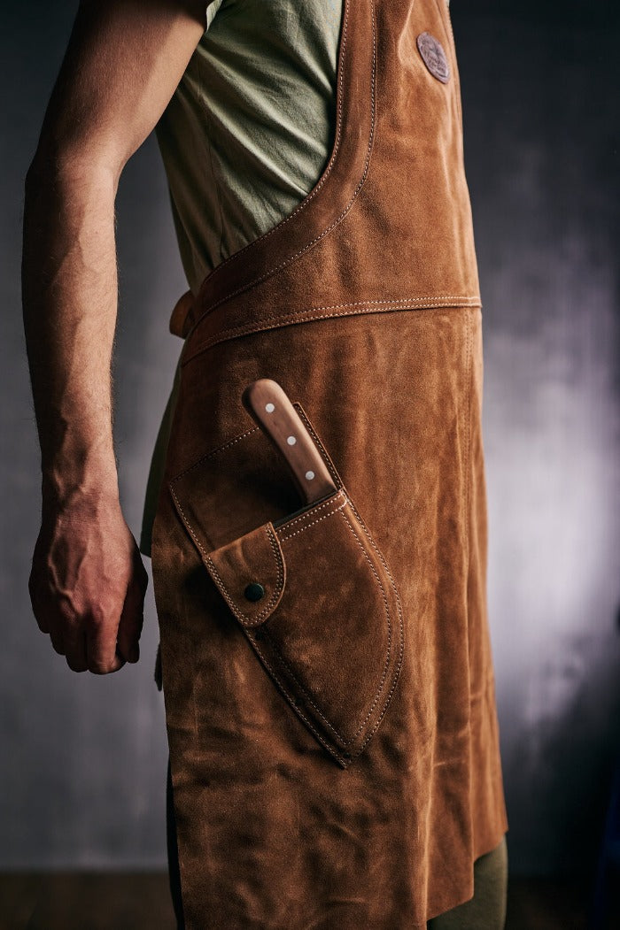 Osoba nosi Almazan Kitchen kožnu kuvarsku kecelju i ima nož u posebnom džepu. 